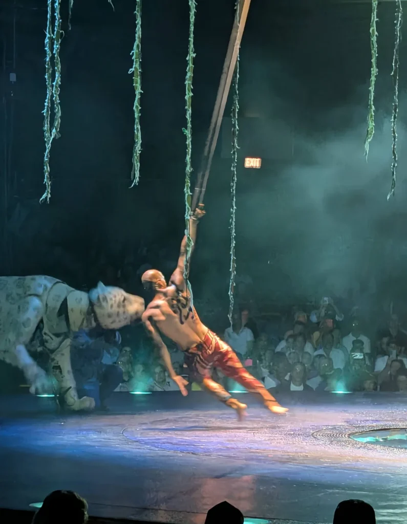 Performer suspenso em fitas aéreas ao lado do puppet da onça-pintada, durante o Cirque du Soleil LUZIA em Miami. Foto própria, 2026.