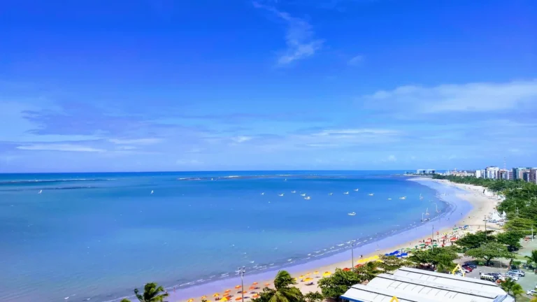 Praia urbana de Maceió com mar verde e linha de recifes ao fundo
