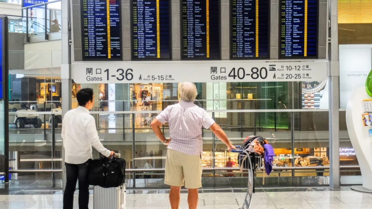Dois viajantes diante do painel de partidas de um aeroporto, avaliando as opções de voo.