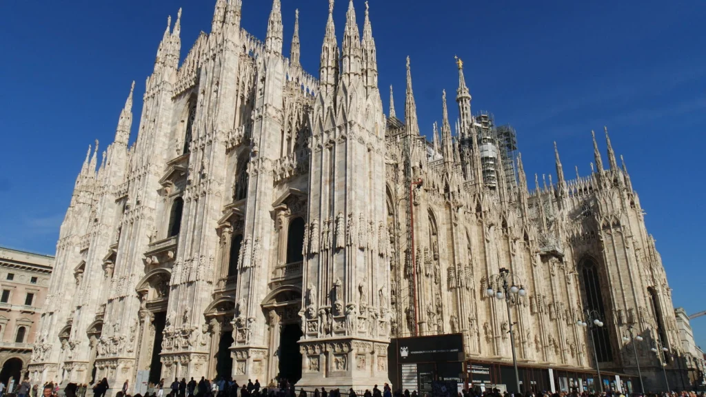 A fachada gótica do Duomo de Milão sob céu azul, na Piazza del Duomo