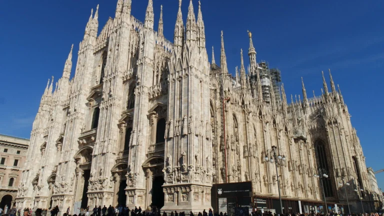 A fachada gótica do Duomo de Milão sob céu azul, na Piazza del Duomo