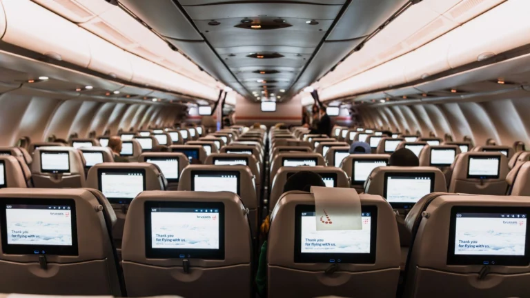 Interior de cabine de avião com fileiras de poltronas e telas de entretenimento individuais em cada encosto, iluminação ambiente suave.