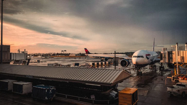 Aviões de fuselagem larga estacionados nos portões de um aeroporto ao entardecer, com pontes de embarque e pista molhada.