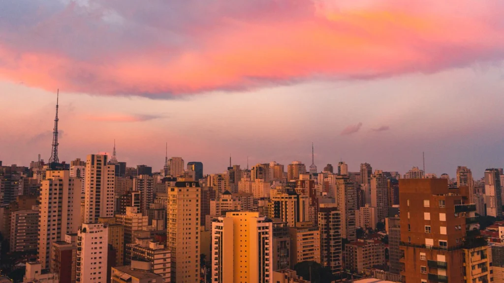 Vista do skyline de São Paulo ao entardecer, com o céu em tons de rosa e laranja sobre os arranha-céus