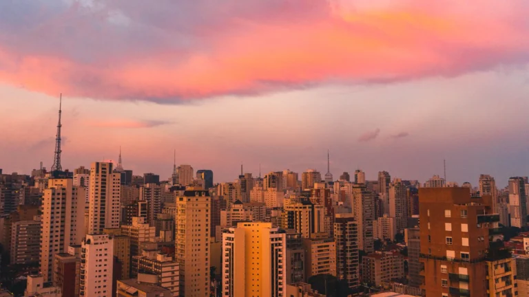 Vista do skyline de São Paulo ao entardecer, com o céu em tons de rosa e laranja sobre os arranha-céus