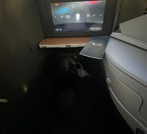 Assento de classe executiva da American Airlines visto do lugar do passageiro, com tela individual, mesa de madeira e espaço para as pernas