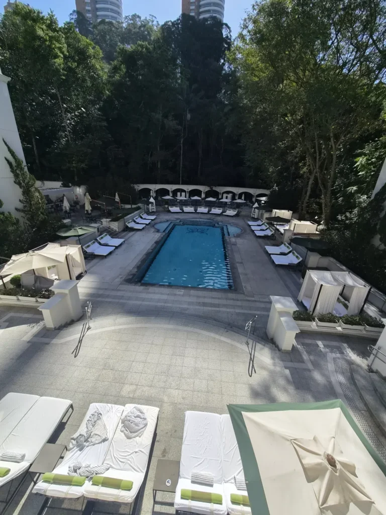 Piscina externa do Palácio Tangará cercada pelo verde do Parque Burle Marx, com camas de sol e cabanas brancas. Foto própria, 2026.